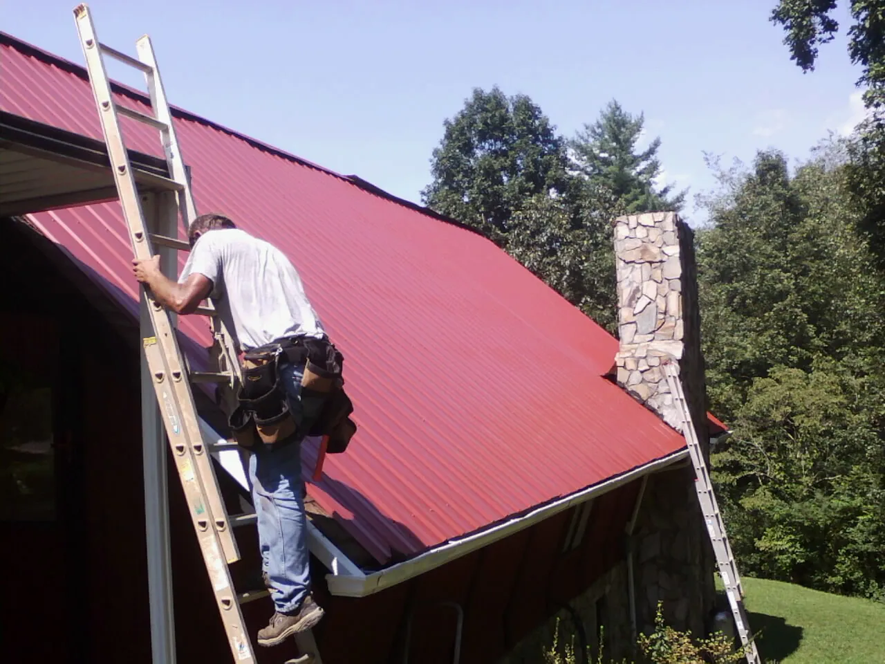 Roofer on a ladder beside a red metal roof and stone chimney — Soffit & Fascia Repair by Roofing Brothers in Sheboygan